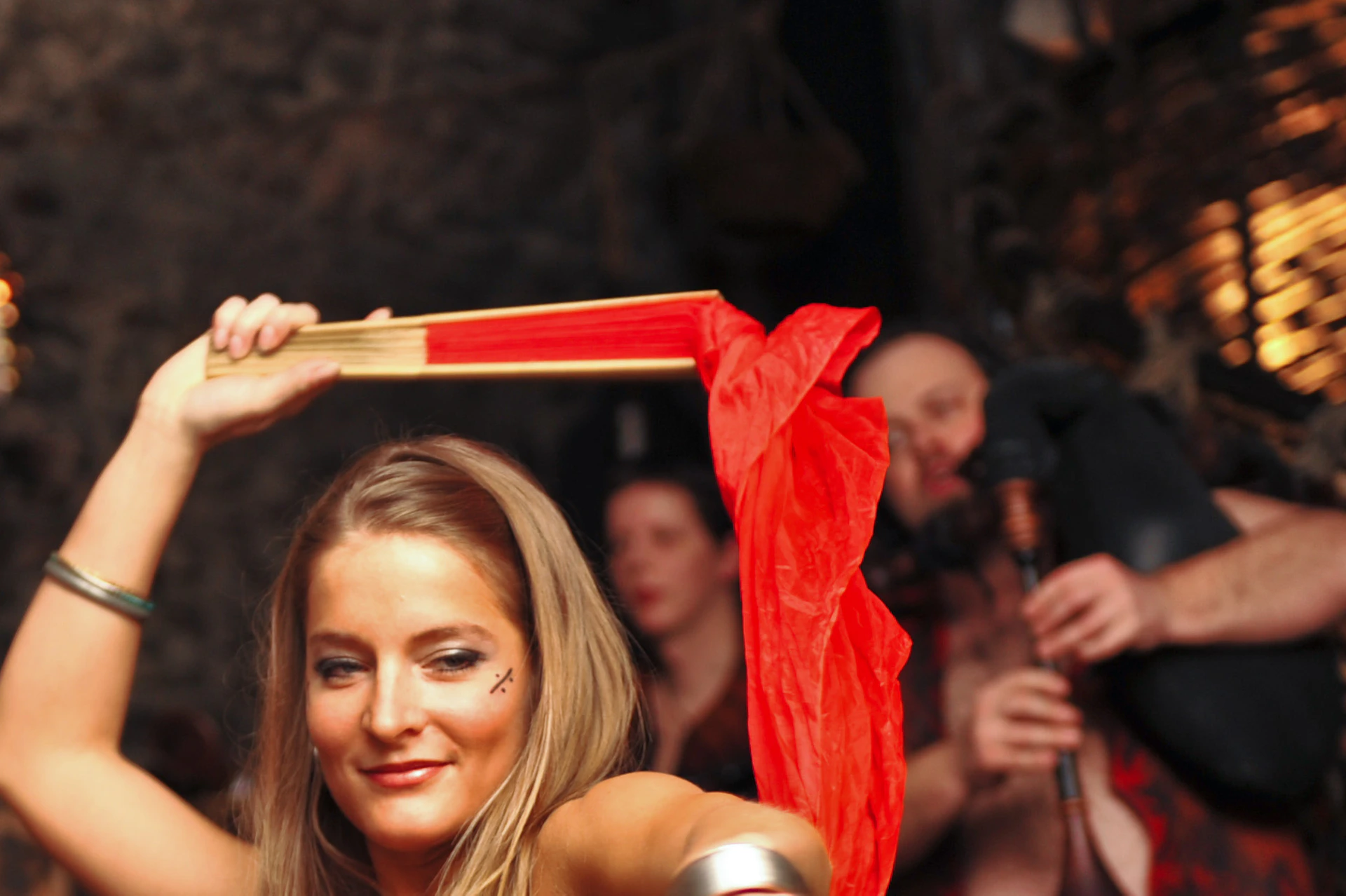 A woman in belly dance attire dances with a red veil in a dimly lit room, while musicians play in the background.