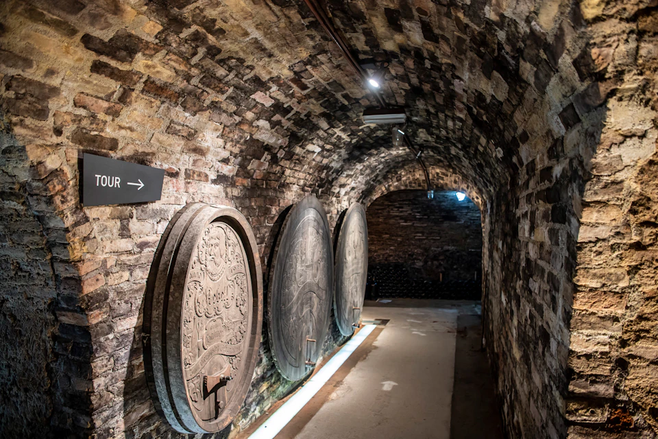 Brick cellar with large, ornate wooden barrels lining the walls and a sign indicating a tour direction. Dimly lit with spotlights.