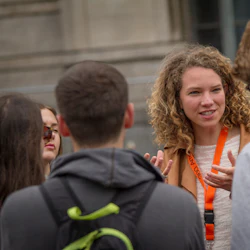 En kvinna med lockigt hår talar till en grupp människor utomhus. Hon bär en orange snodd och gestikulerar med händerna.