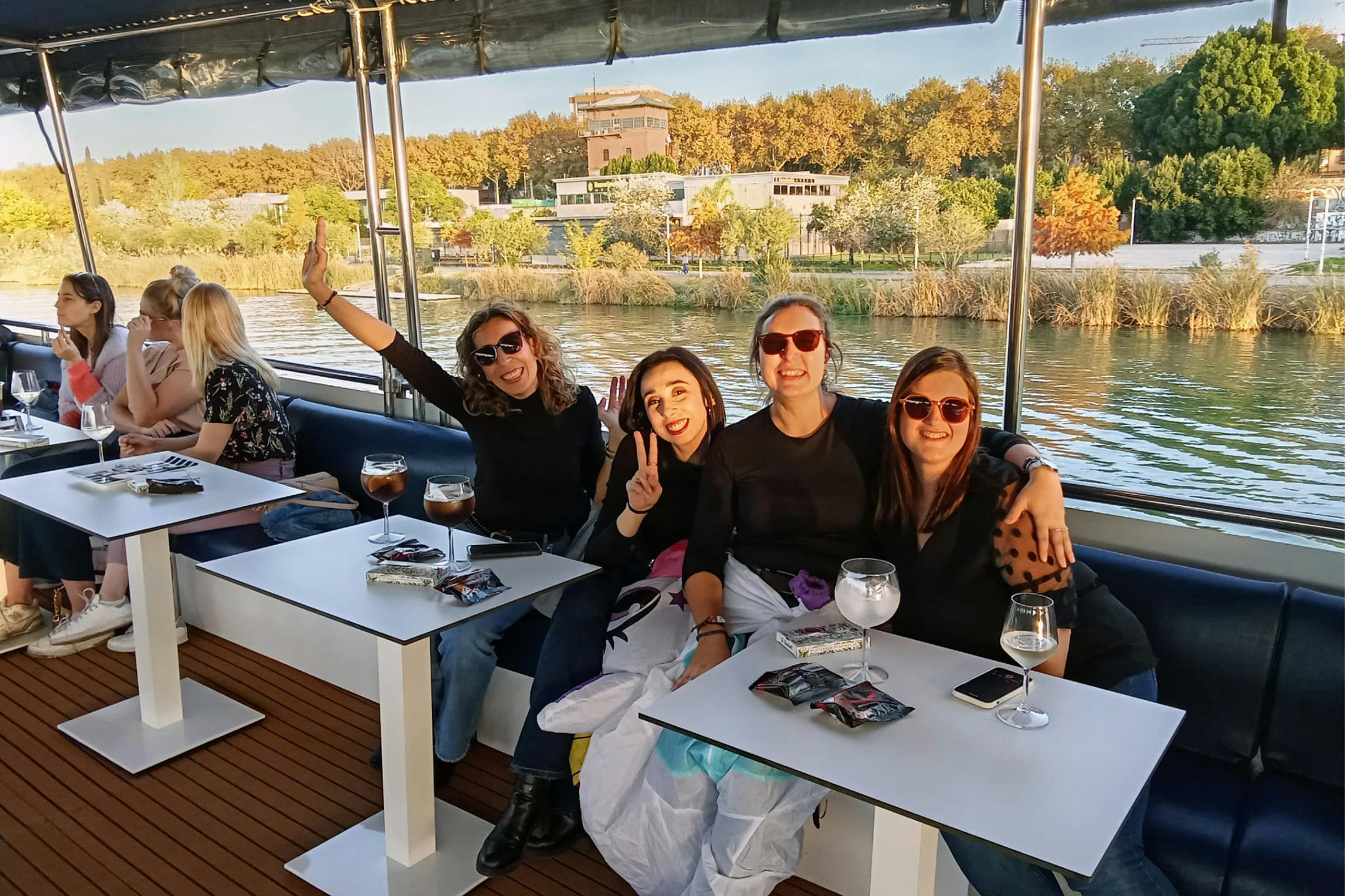 Cztery kobiety na łodzi nad brzegiem rzeki, uśmiechające się i pozujące przy białym stole z napojami i telefonami.