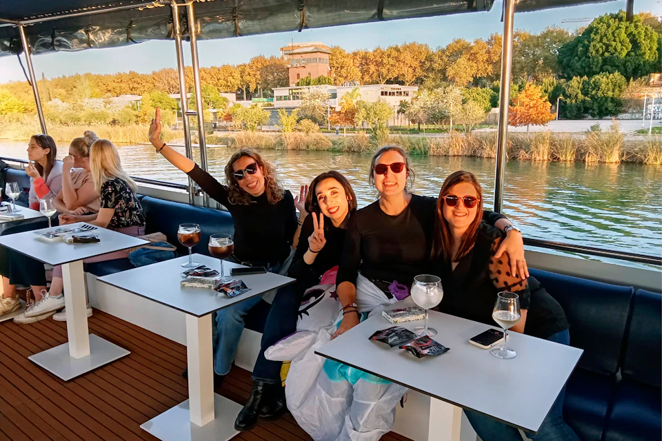 Four women on a boat near a riverbank, smiling and posing at a white table with drinks and phones on it.