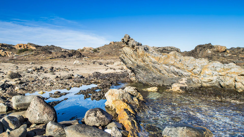 Rocky coastal landscape with tide pools, large boulders, and clear blue sky.