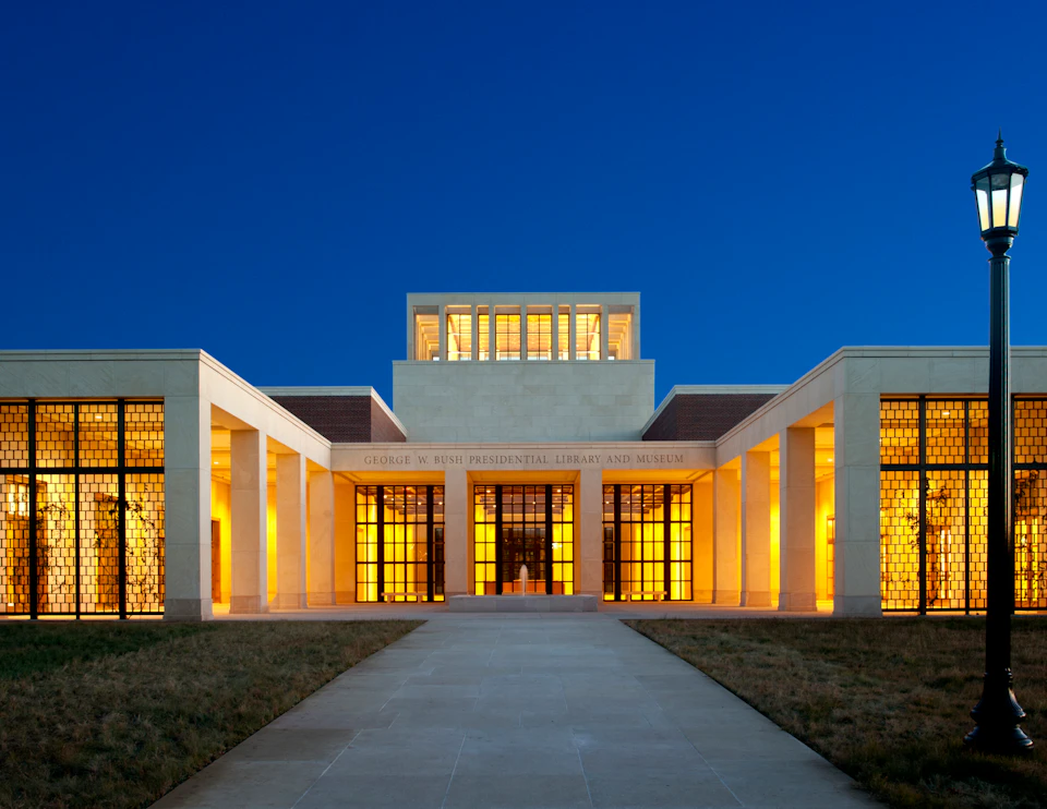 Illuminated entrance of the George W. Bush Presidential Library and Museum at dusk, featuring modern architecture and glass doors.