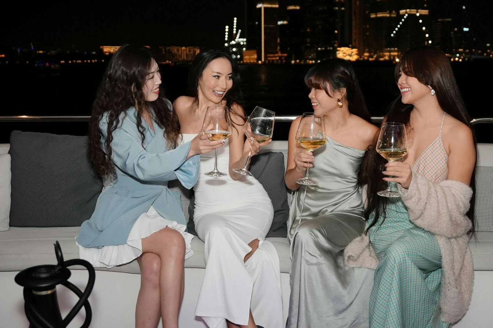Four women seated outdoors, smiling and holding wine glasses, with a cityscape illuminated at night in the background.