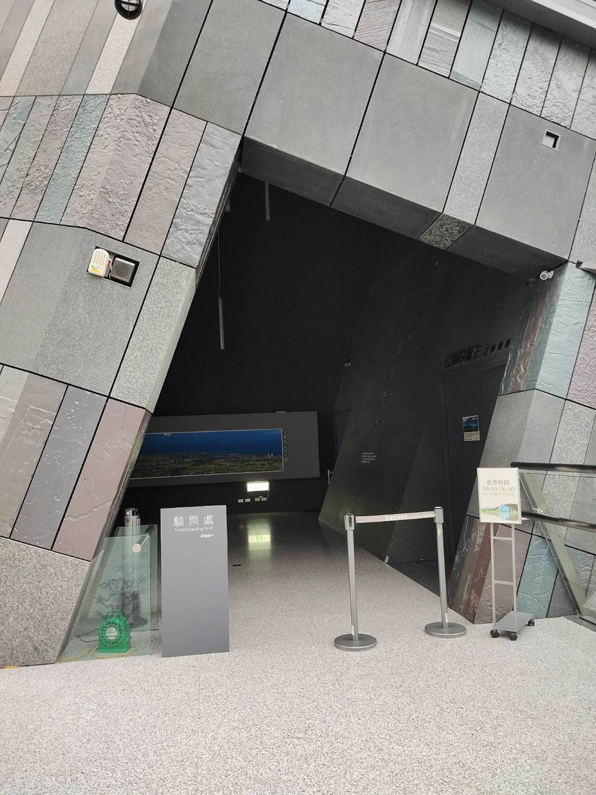Entrance to a modern building with dark geometric walls, a rope barrier, and informational signage near the doorway.