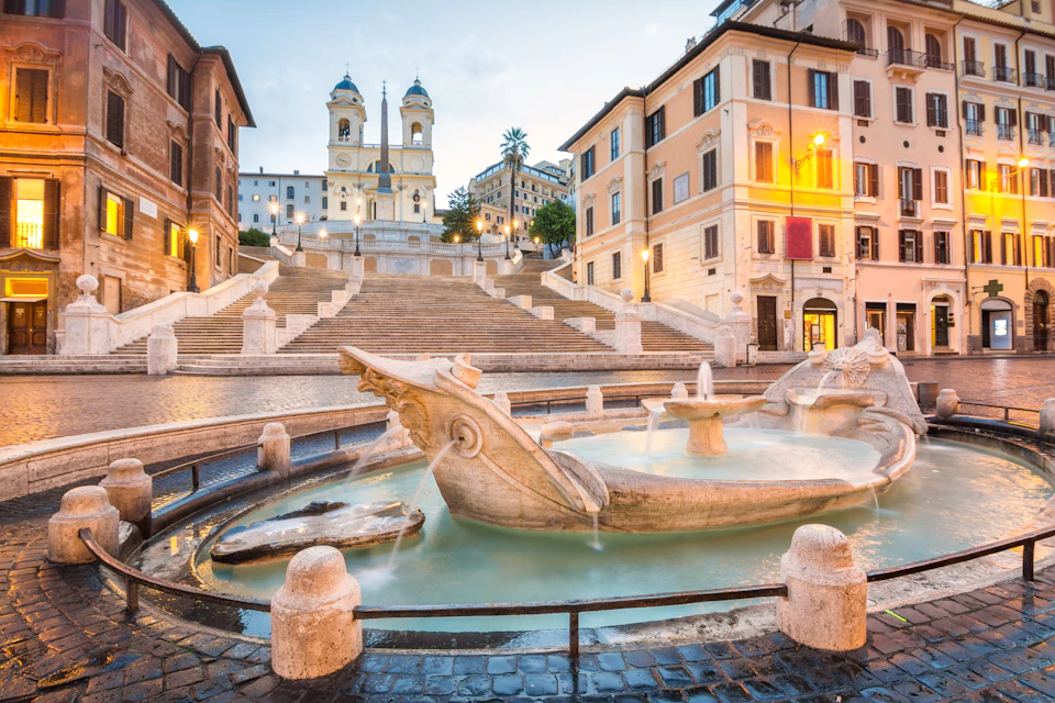 An ornate fountain sits in front of the Spanish Steps, leading up to the Trinità dei Monti church in Rome at dawn.
