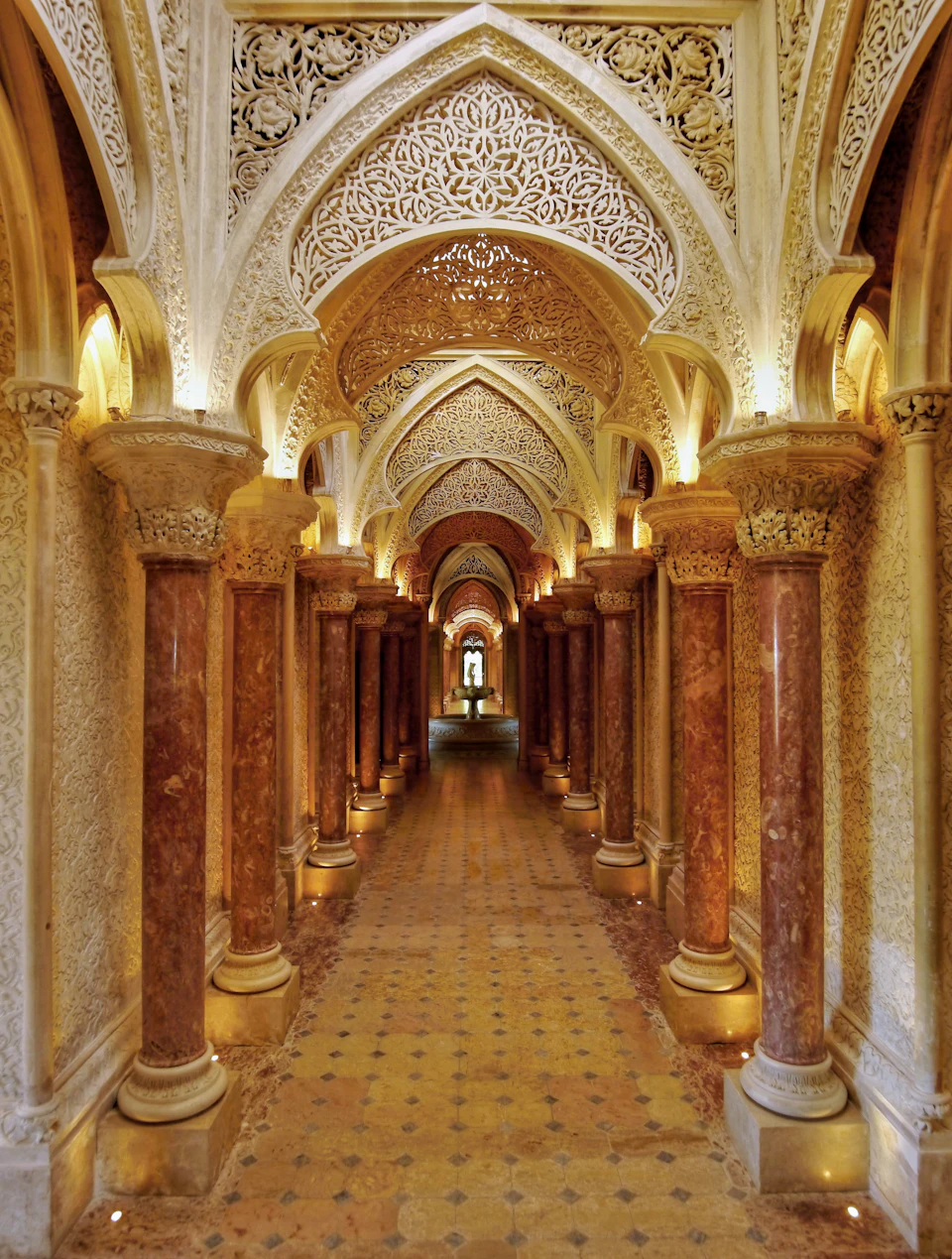 Ornate hallway with intricate arches, decorative carvings, and red marble columns leading to a light-filled area.