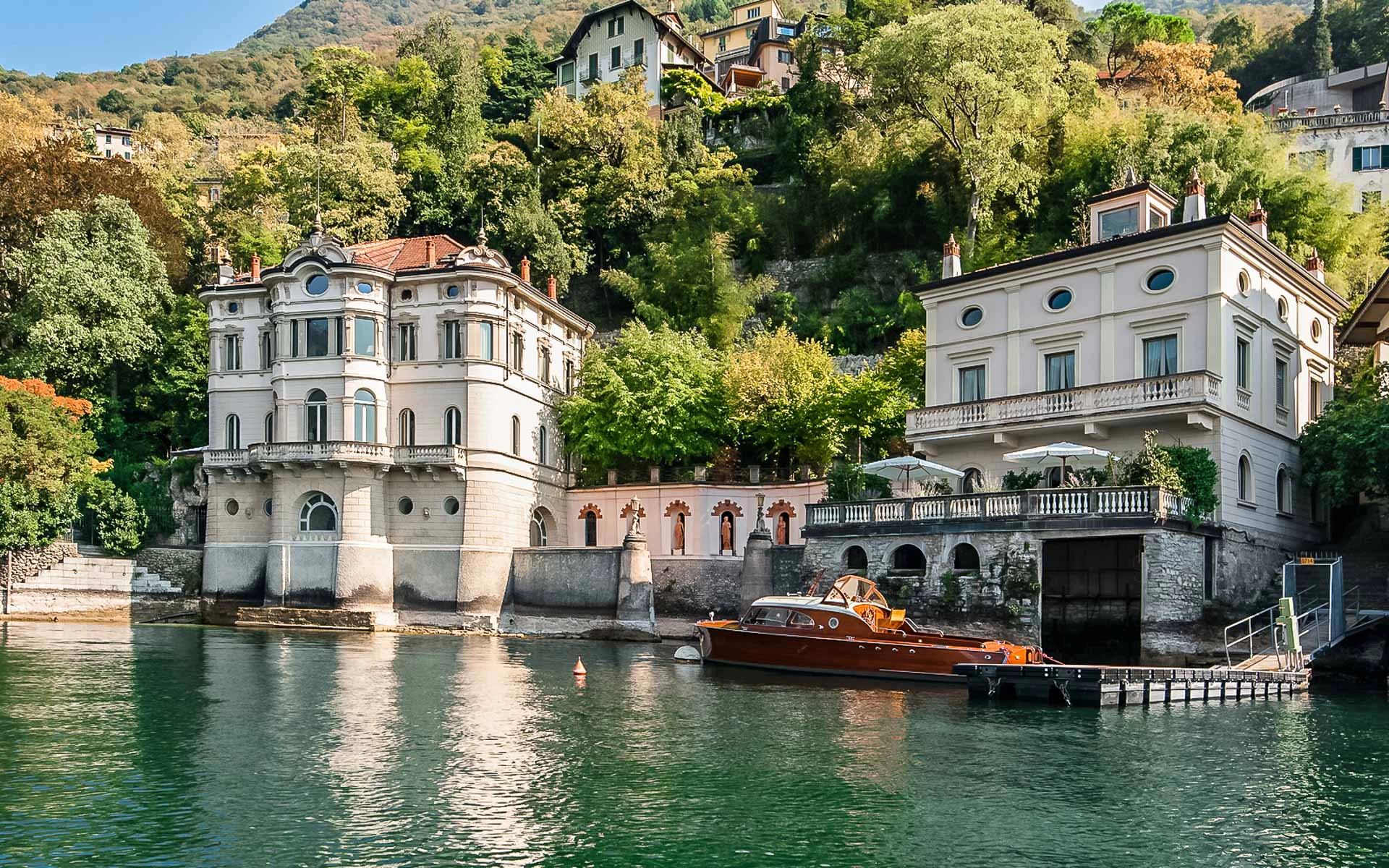 Large, elegant waterfront houses with arched windows and balconies, nestled against a lush, green hillside, with a boat docked in front.