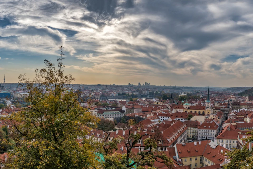 Aerial view of a city with red-tiled rooftops, trees in the foreground, and dramatic cloud patterns in the sky.