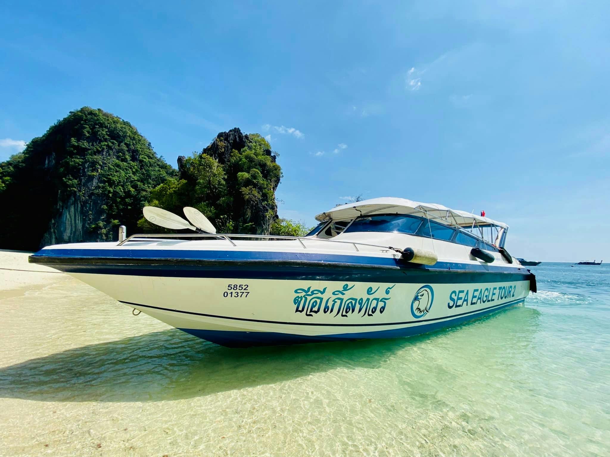 Ένα ταχύπλοο με την ένδειξη "SEA EAGLE TOUR 1" είναι αγκυροβολημένο σε καθαρά ρηχά νερά κοντά σε βραχώδεις, δεντροσκεπείς γκρεμούς κάτω από έναν καταγάλανο ουρανό.