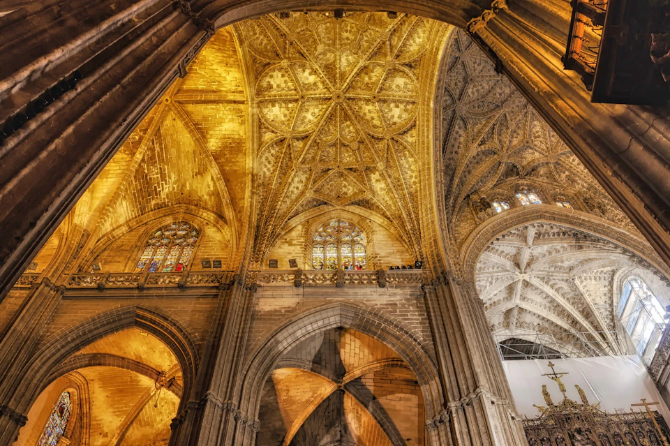 Ornate Gothic cathedral ceiling with pointed arches, intricate gold detailing, and colorful stained glass windows.