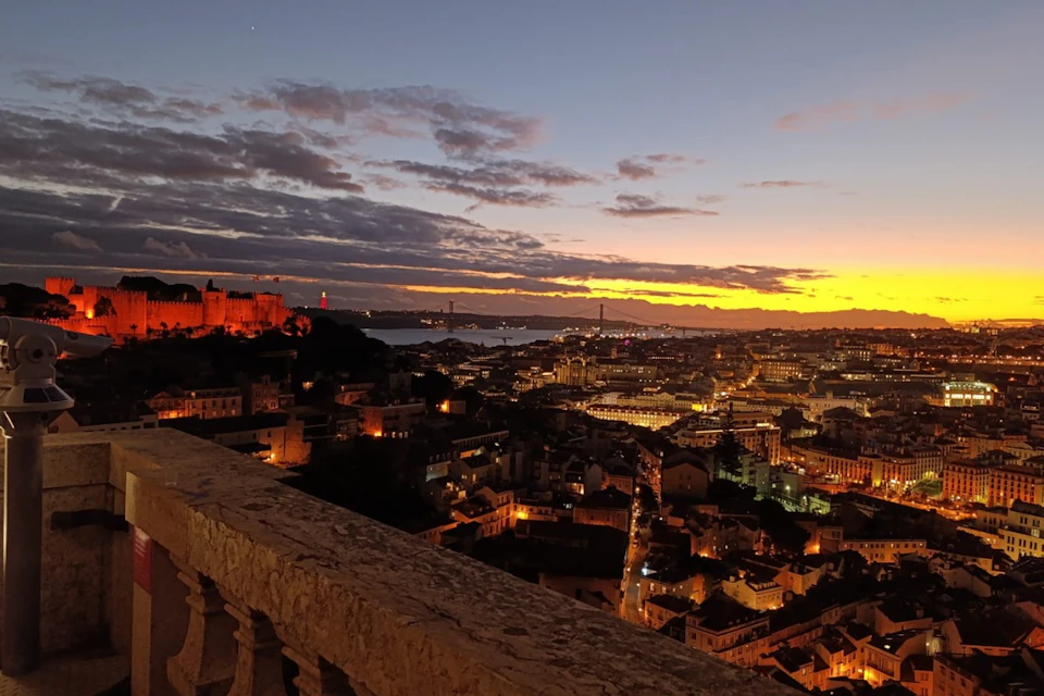 A cityscape at sunset with buildings illuminated, a distant bridge, and a castle on a hill, viewed from behind a stone railing.