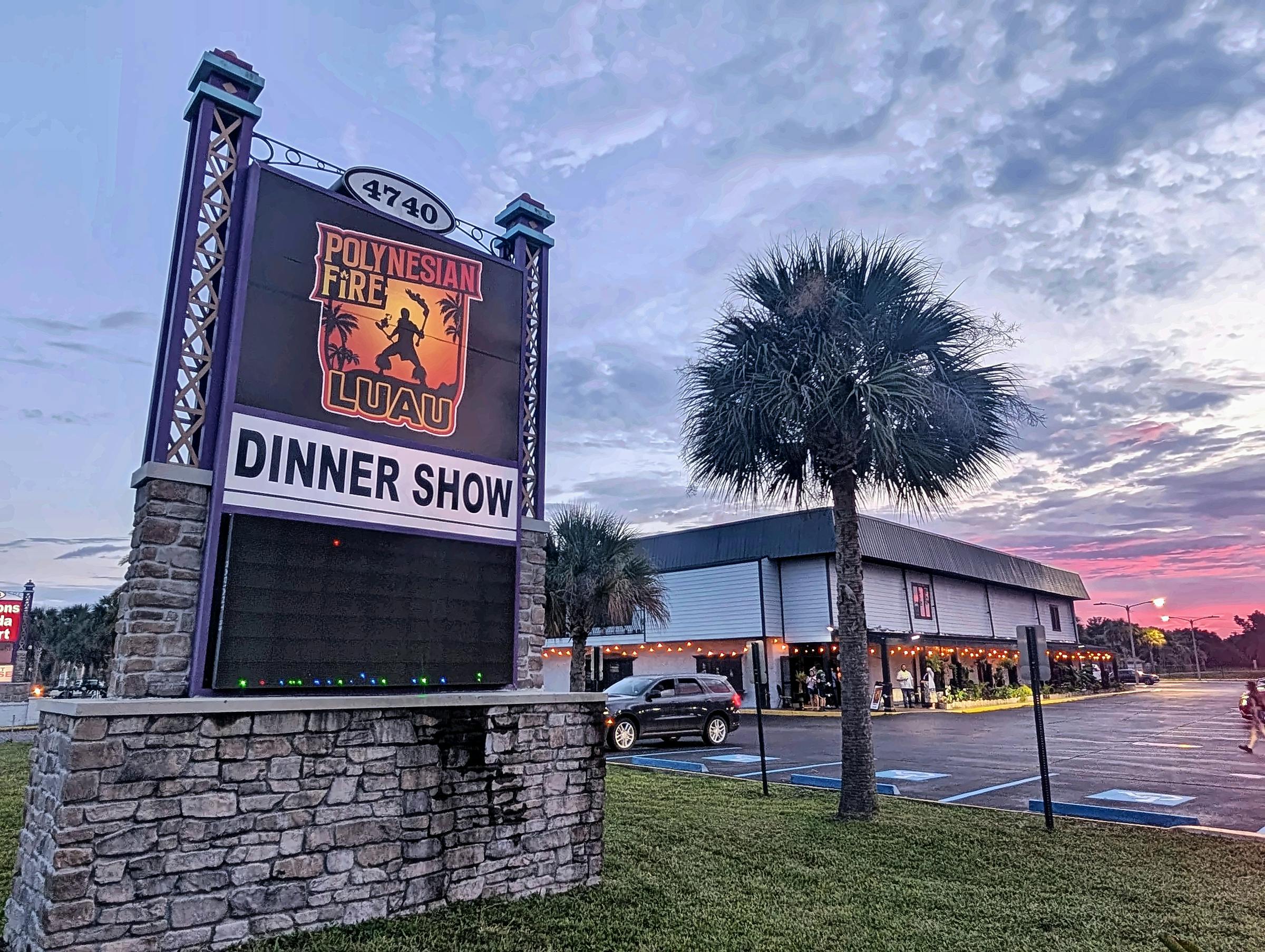 A sign reading 'Polynesian Fire Luau Dinner Show' with a large building, palm trees, and parked cars in the background.