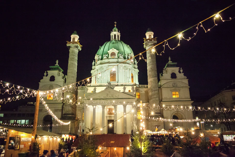 Illuminated baroque church with a green dome and twin columns at night, with festive string lights and a crowded market below.