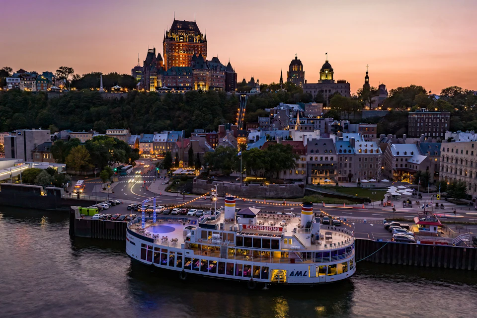A ship docked by a waterfront with a historic cityscape in the background at sunset, including a castle-like building.