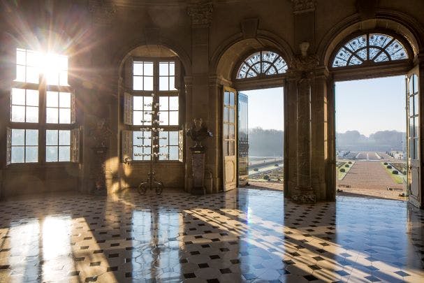Sunlight streaming through large arched windows and doors in an elegant hall with a tiled floor and outdoor view.