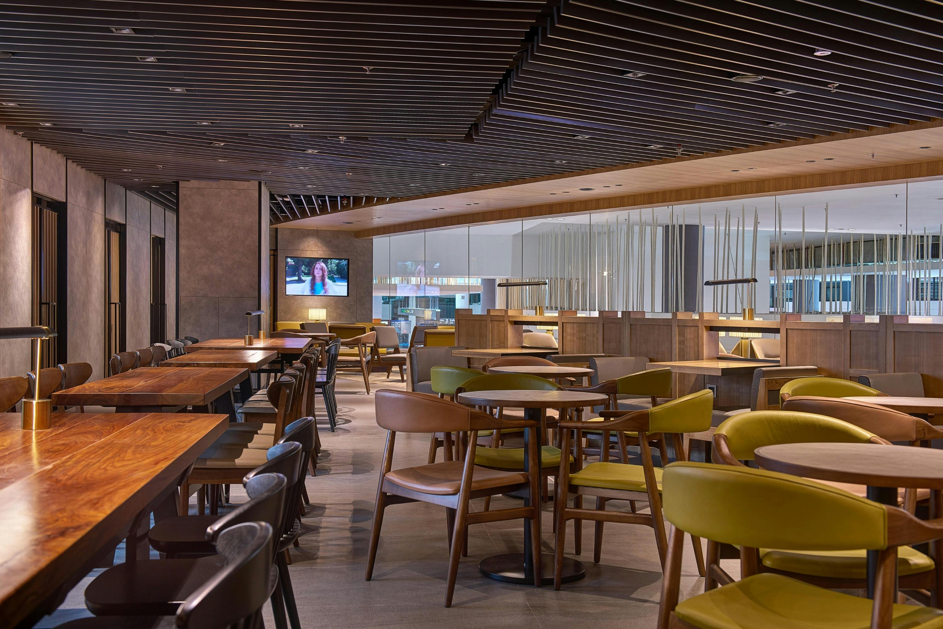A modern restaurant interior with wooden tables, chairs, and a TV screen on the wall.