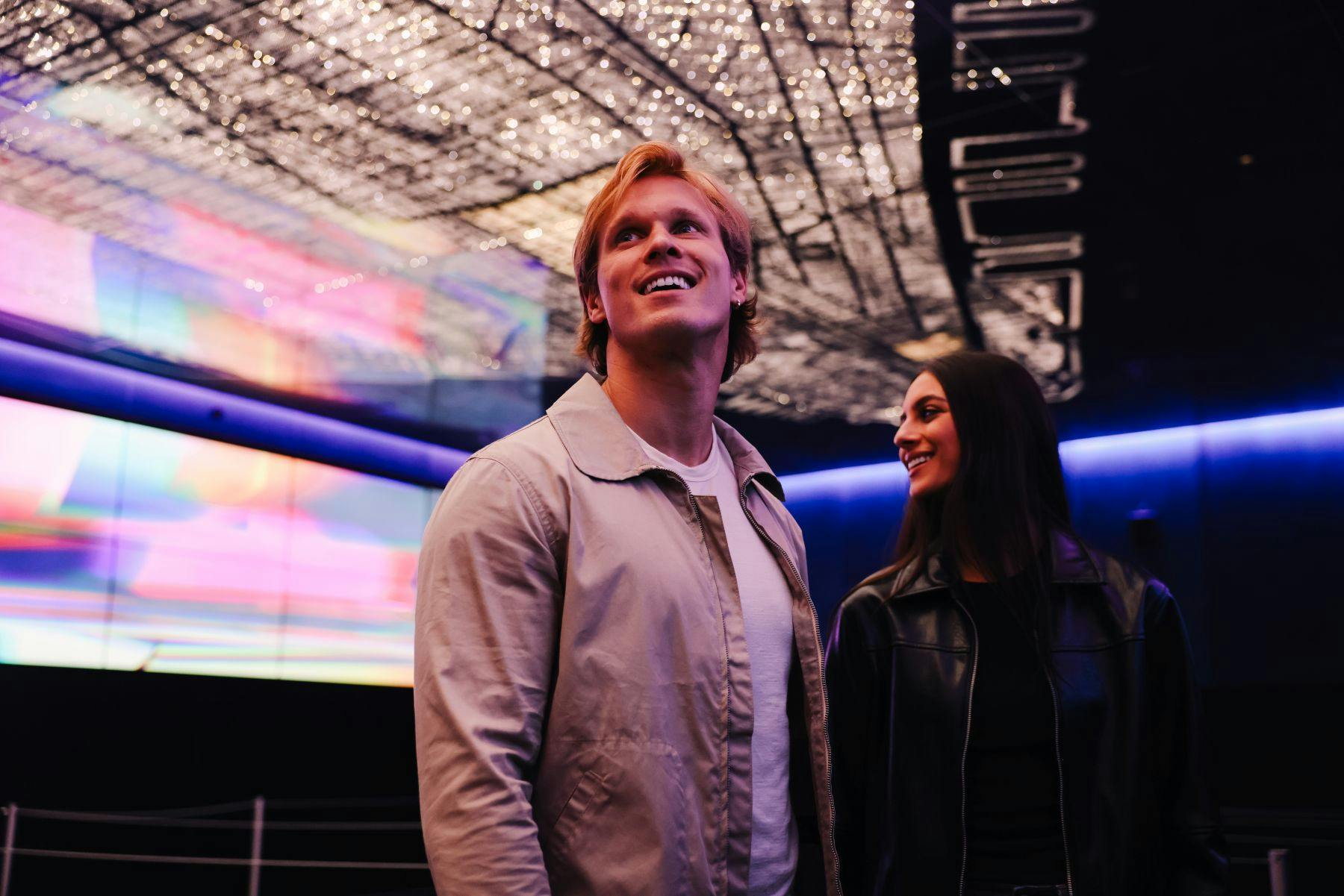 Two people standing indoors, looking up and smiling, with a vibrant, illuminated ceiling in the background.