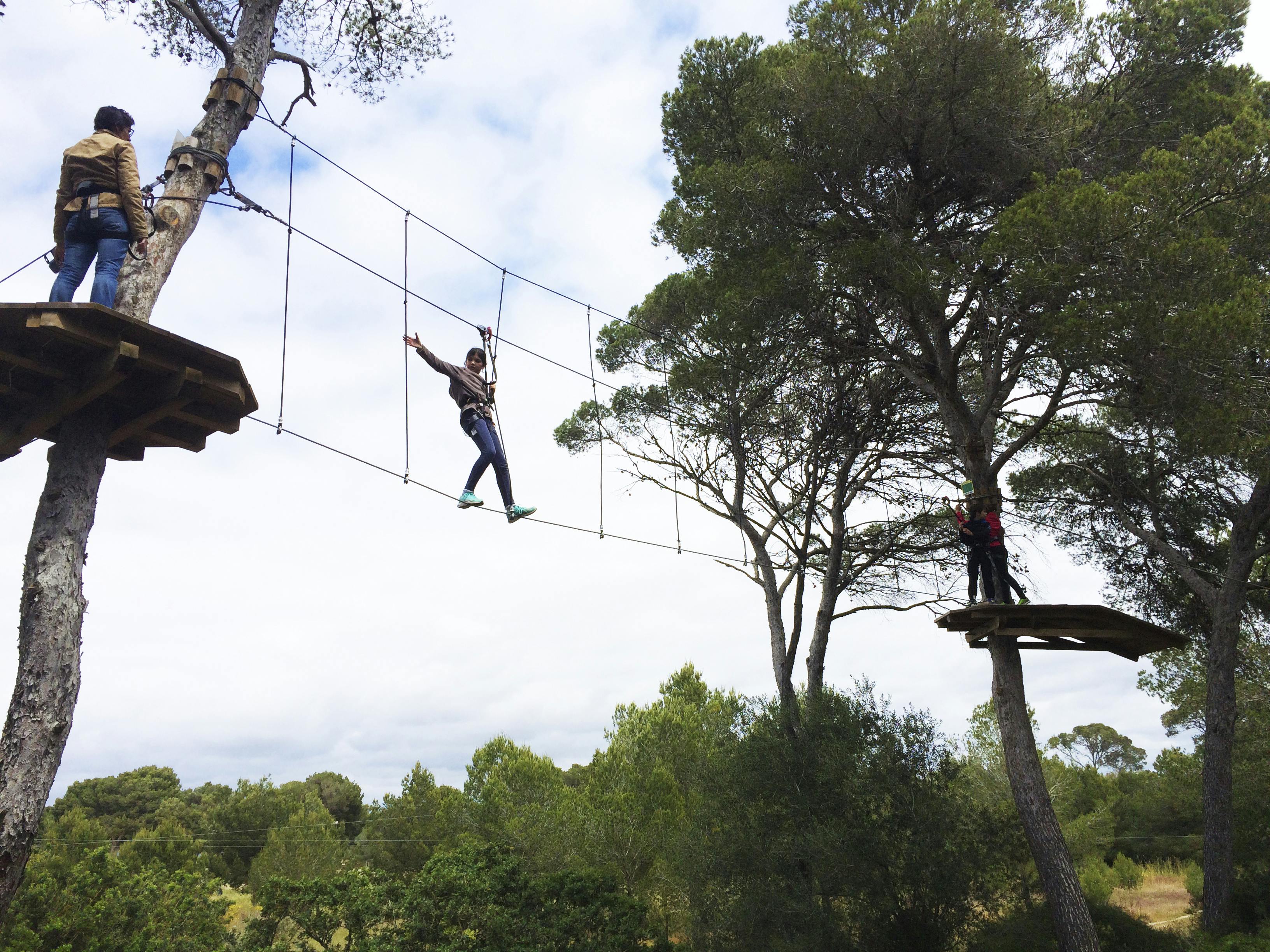 Person walking on a rope bridge between trees in a forest, with another person standing on a wooden platform nearby.