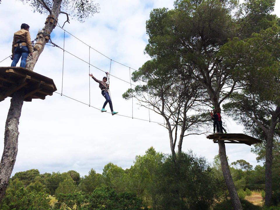Person walking on a rope bridge between trees in a forest, with another person standing on a wooden platform nearby.