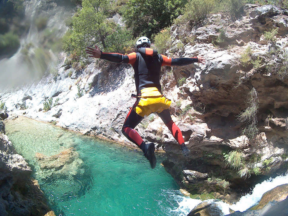 Person in a wetsuit and helmet jumping off a rocky ledge into a clear turquoise pool below.