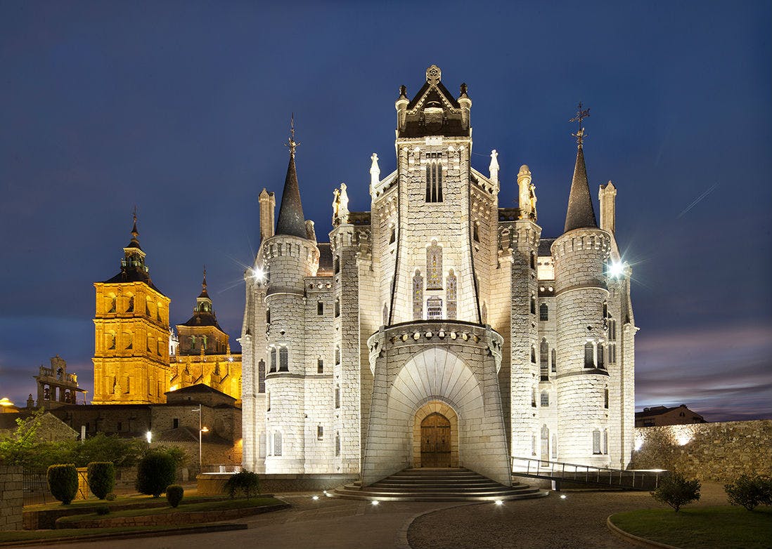 Illuminated, ornate castle with turrets and arch entrance at dusk, with a historic church in the background.
