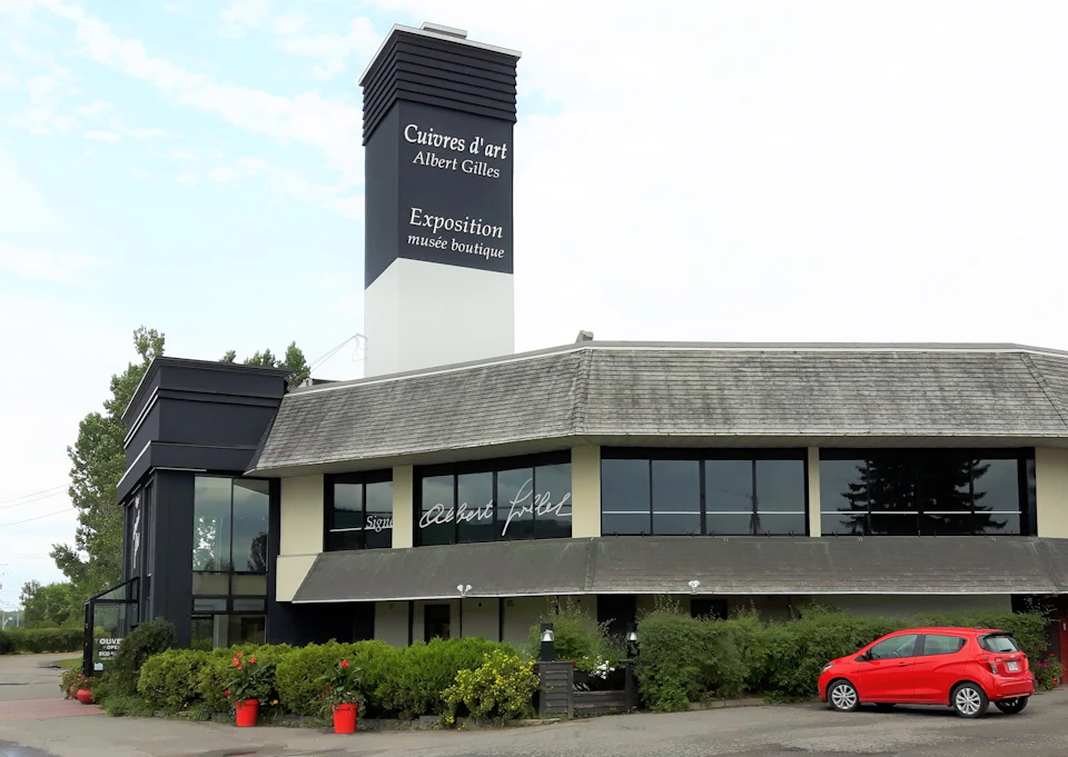 A building with a tall sign that reads "Cuivres d'art Albert Gilles," featuring a museum boutique. Bushes and red car in front.