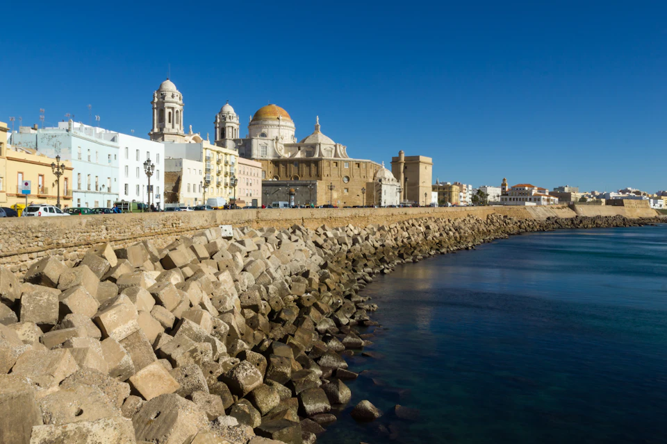 Historic buildings with domes and towers line a rocky coastal promenade under a clear blue sky.