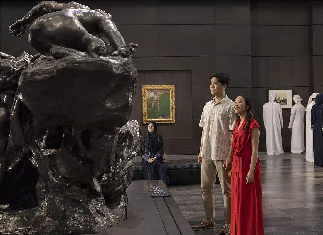 A man and woman observe a large sculpture in an art gallery, surrounded by other visitors and framed paintings.