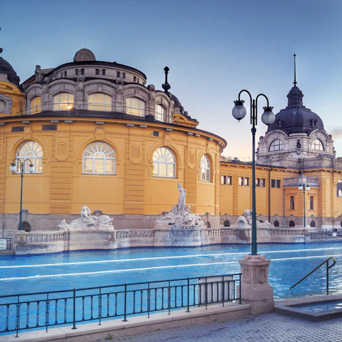 Utsmyckad gul byggnad med välvda fönster, kupolformat tak, statyer och en pool i förgrunden under en klar himmel.