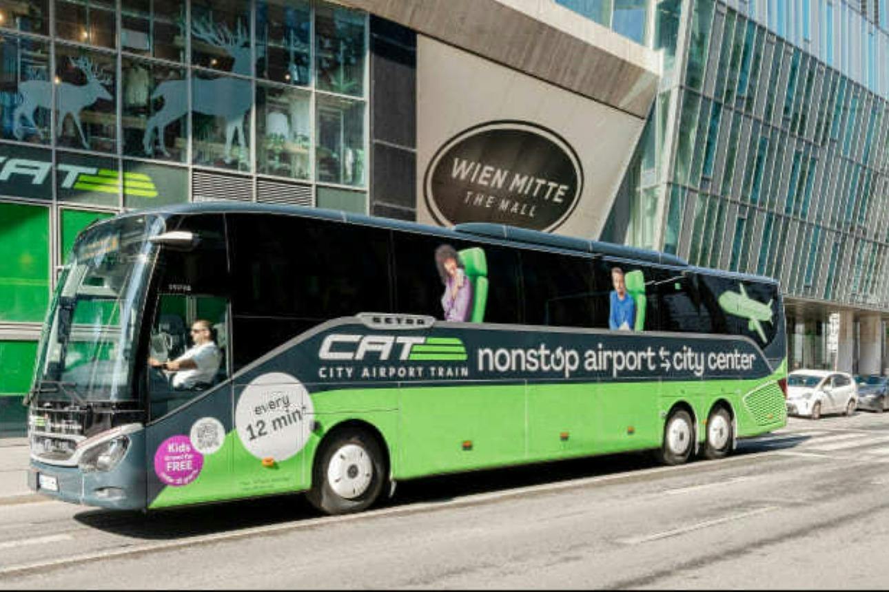 En grøn og sort bybus mærket 'City Airport Train' parkerede foran et indkøbscenter med glasvinduer.