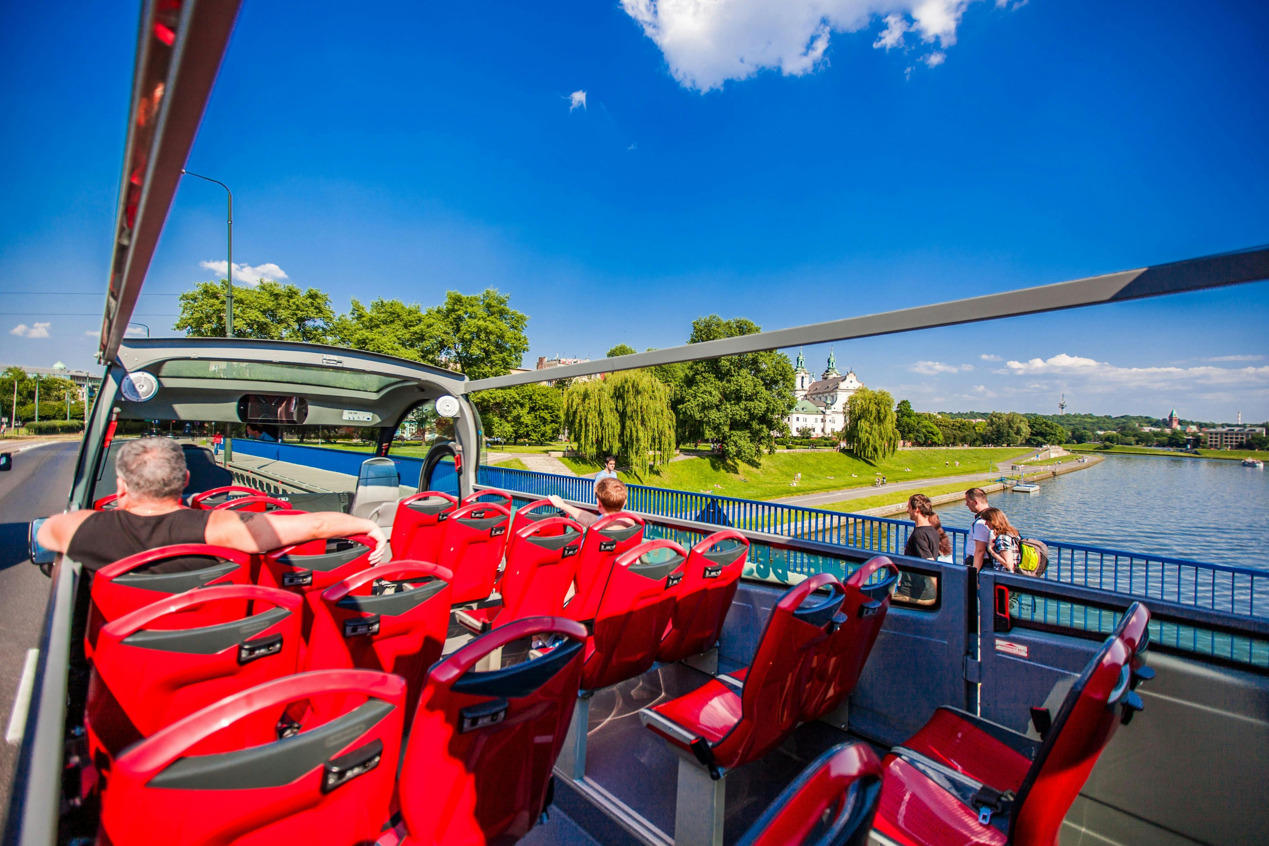 Open-top bus with red seats, people seated, passing a river with lush greenery and a white church in the background under a clear blue sky.