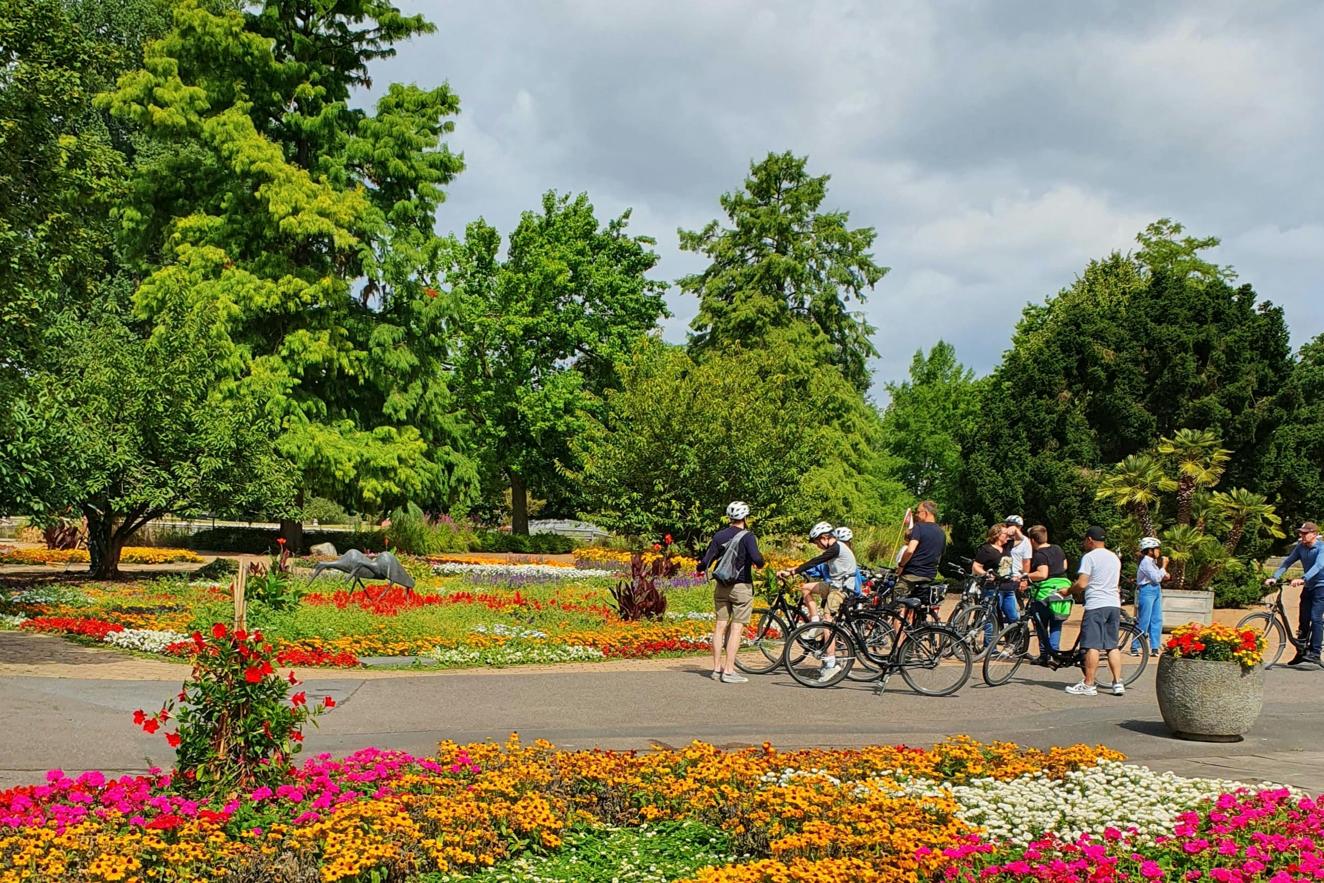 Skupina lidí s jízdními koly se shromáždí na pěšině v barevné zahradě se zářivými květinovými záhony a vysokými zelenými stromy.