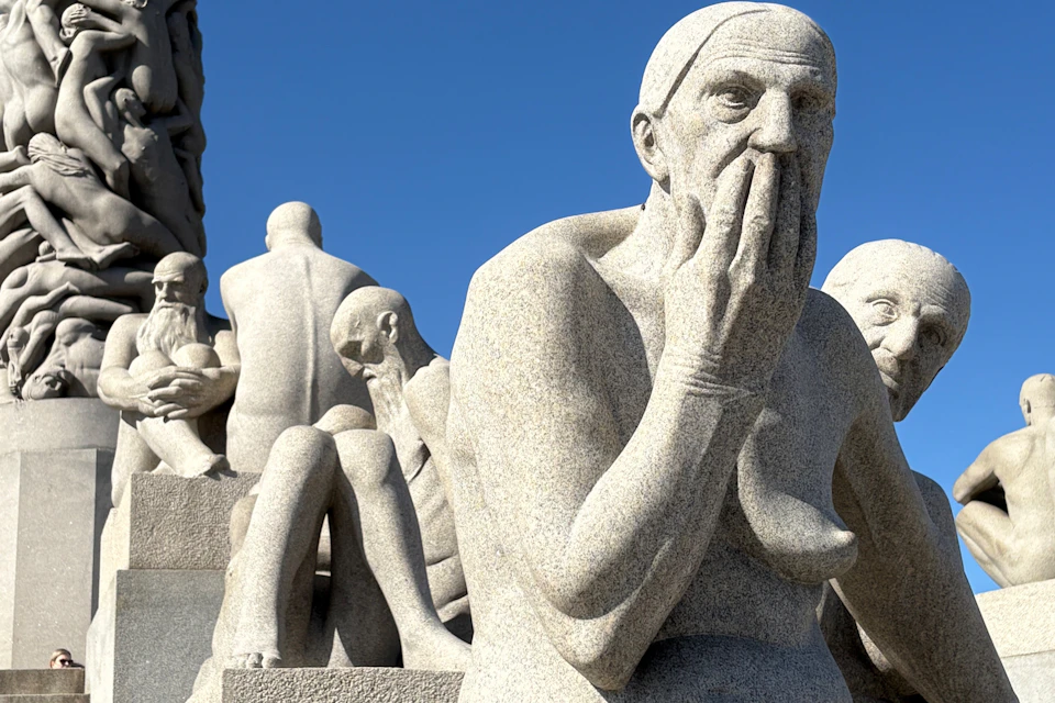 Multiple stone sculptures of human figures are seated closely together, with one figure covering its mouth. The sky is clear and blue.