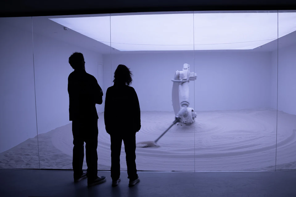 Two people observe a robotic arm creating patterns in sand inside a glass-enclosed room.