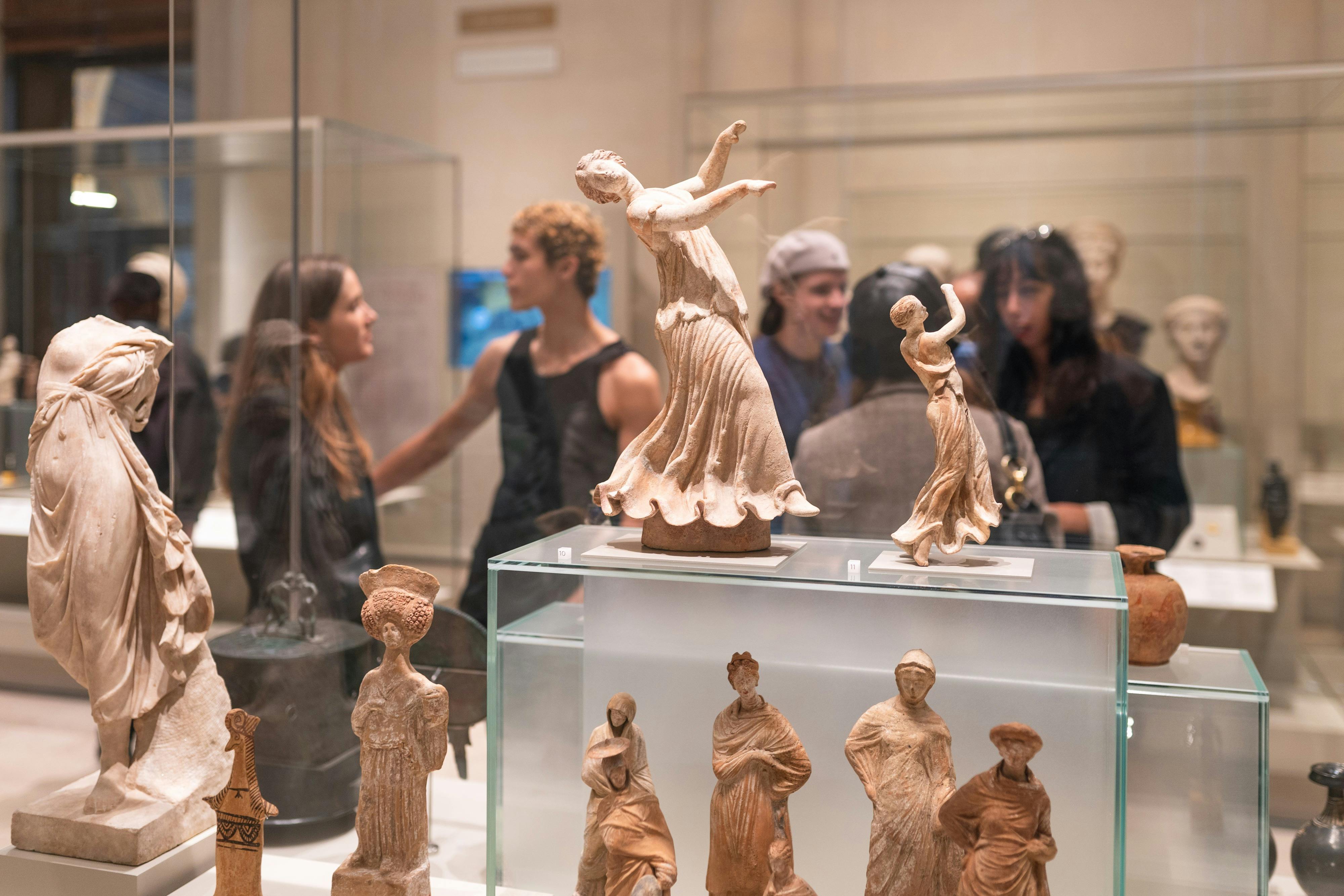 An exhibition showcasing ancient figurines on glass displays with visitors observing in the background.