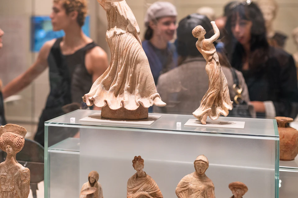 An exhibition showcasing ancient figurines on glass displays with visitors observing in the background.
