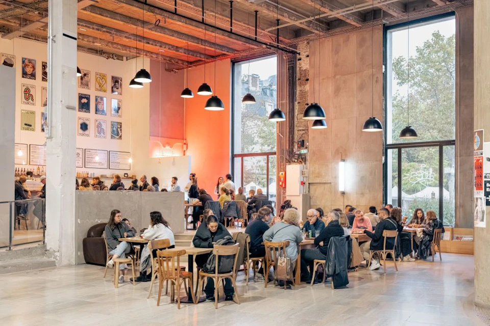 A lively cafe with people seated at tables, large windows, pendant lights, and modern industrial decor.