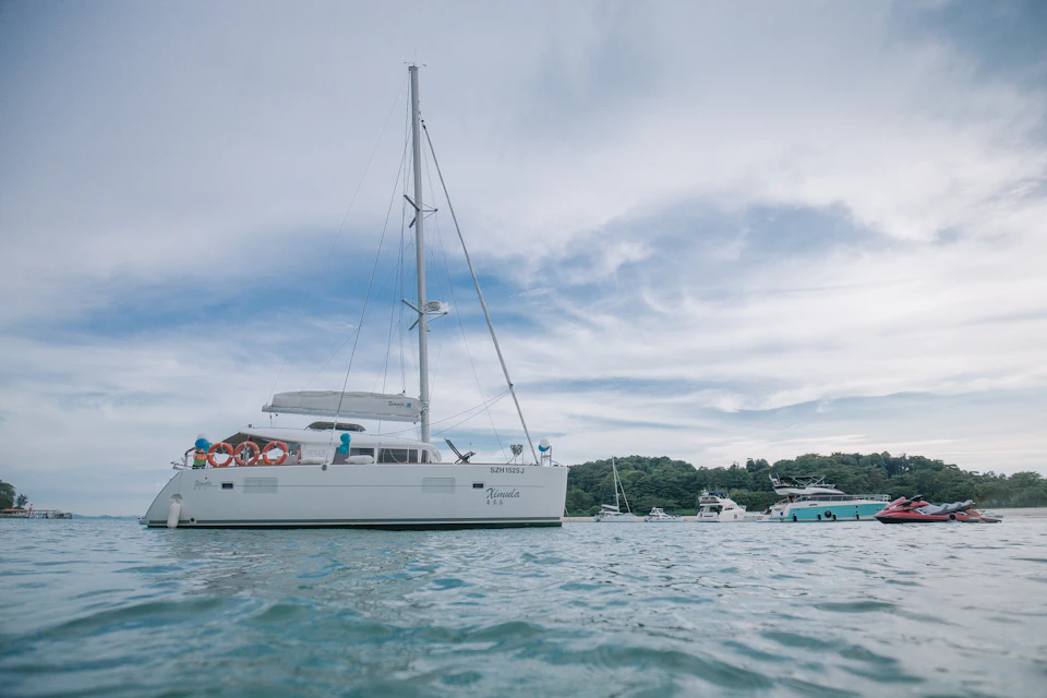 A white sailboat on calm water with trees and other boats in the background under a partly cloudy sky.