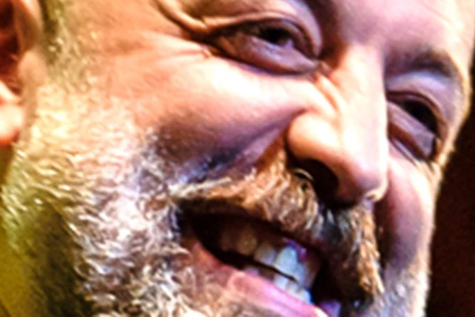 A person holding a microphone smiling on stage with warm lighting and a blurred background.