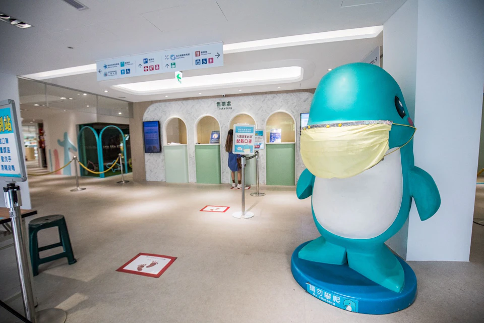 A reception area with service counters, a person standing in line, and a large cartoon figure wearing a mask in the foreground.
