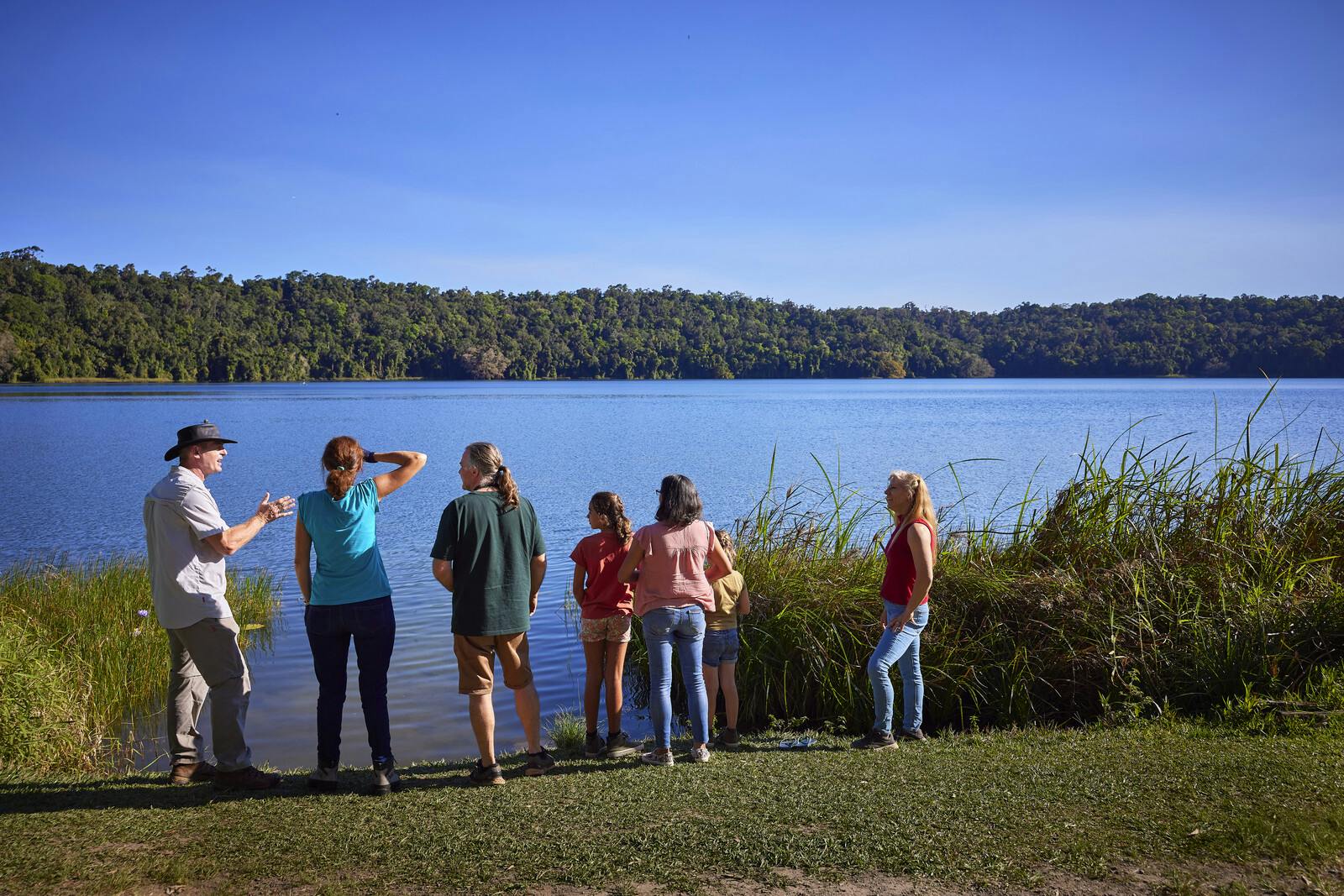 澄んだ青い空の下、木々を背景に、草むらの湖畔の近くに立っている人々のグループ。
