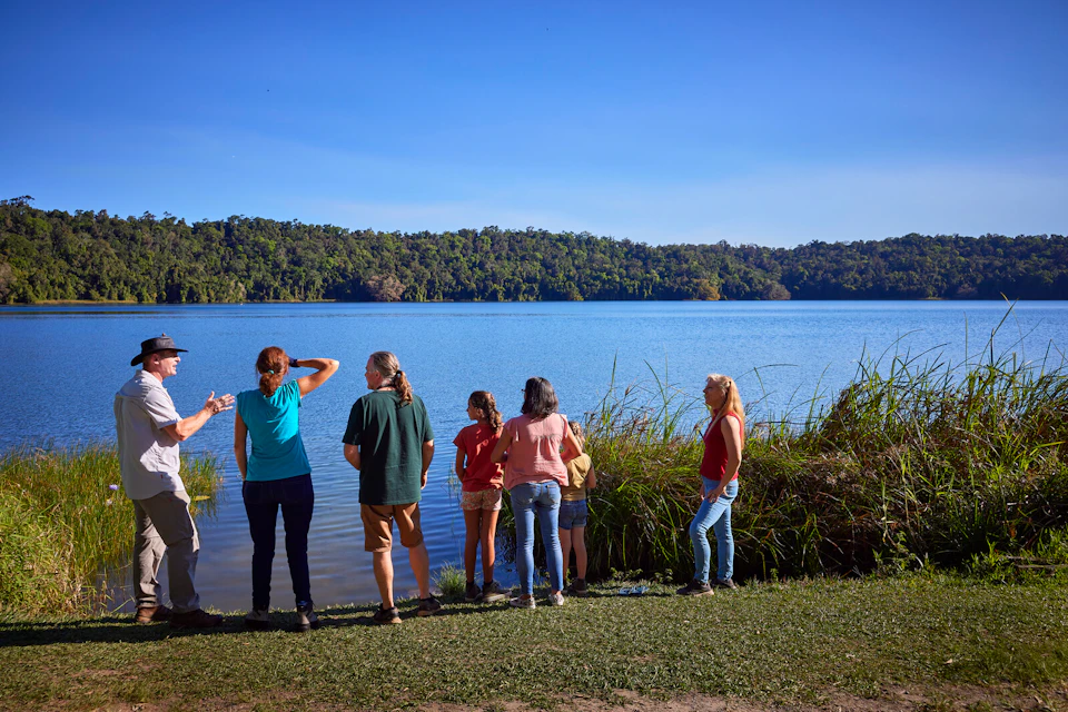 A group of people standing near a grassy lakeside with trees in the background under a clear blue sky.