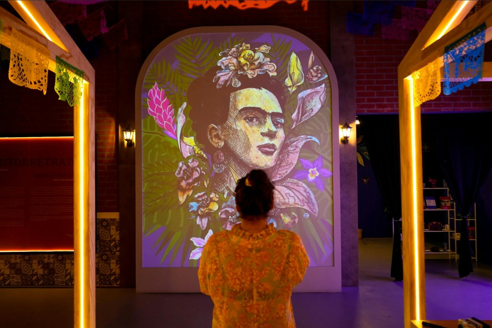 Πρόσωπο που παρακολουθεί μια μεγάλη, πολύχρωμη καλλιτεχνική προβολή μιας γυναίκας που περιβάλλεται από λουλούδια και φύλλα σε έναν εσωτερικό εκθεσιακό χώρο.