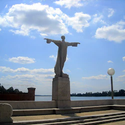 Staty av en figur med utsträckta armar på en piedestal, med utsikt över en flod under en delvis molnig himmel.