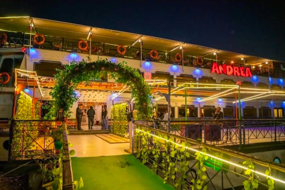 An illuminated entrance to a decorated building at night, with greenery, lights, and people entering the venue.