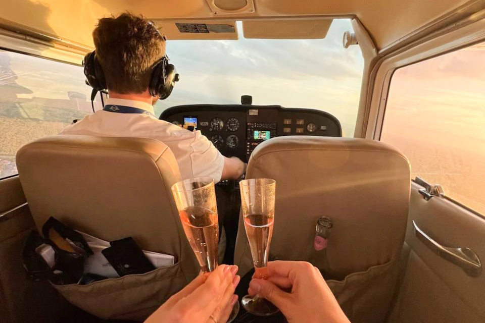 A pilot flying a small aircraft with two hands in the foreground clinking champagne glasses.