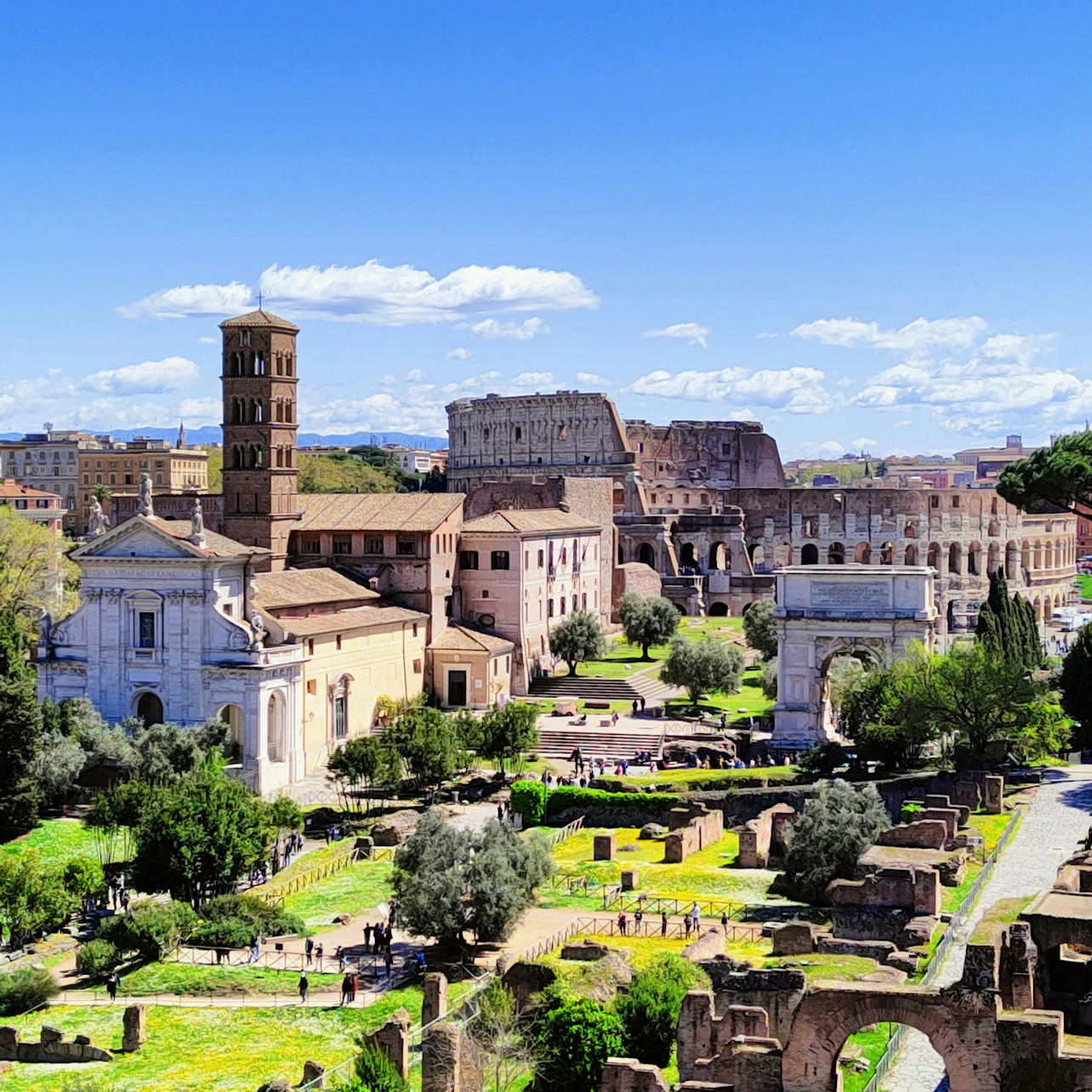 Ancient Roman ruins with the Colosseum, Arch of Constantine, and a bell tower. Greenery and scattered people visiting the site.