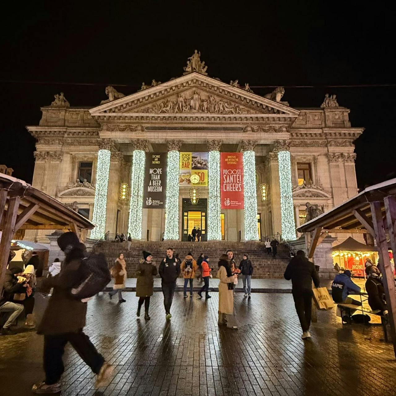Velkolepá, osvětlená budova se sloupy, zdobená pro svátky, obklopená lidmi a slavnostními stánky v noci.