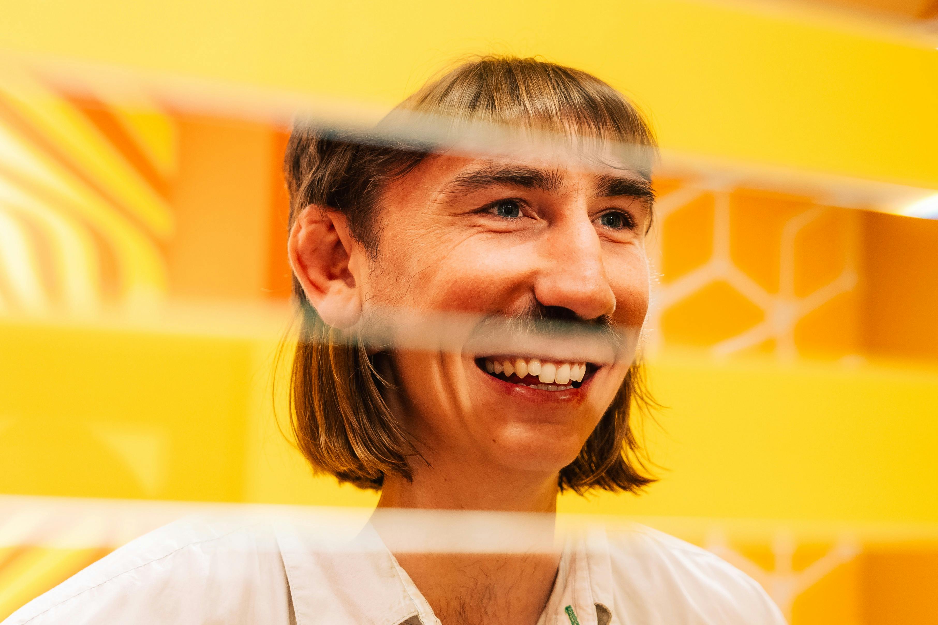 微笑的人，长发肩膀，穿着浅色衬衫，在水平间隙的结构后面，背景是黄色的。